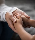 Close up compassionate young foster parent holding hands of little kid girl, giving psychological help, supporting at home. Sincere different generations family sharing secrets or making peace.